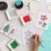 A hand paints Christmas cards featuring holiday designs like a snowman, rocking horse, and gifts. Bowls of red and green paint, tags, and crafting supplies are arranged on a white table.