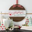 Round sign with Santa and reindeer, “Merry Christmas to All” text, red bow on top. Surrounded by white vases, geometric trees, gold bells, white bead garland, red berries, and greenery on a shelf.