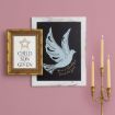 A framed print with a star and text beside a framed dove illustration on a pink wall, next to a gold candelabra holding five lit candles.