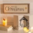 A framed “merry Christmas” sign, a glowing angel candle on books, and a folding nativity scene with Joseph, Mary, and baby Jesus sit on a wooden surface with soft yellow lights.