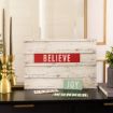 A decorative display features a sign reading “BELIEVE,” with smaller signs below saying “PEACE,” “JOY,” and “WONDER.” The setup includes brass candlesticks and a mirror, suggesting a festive or holiday theme.