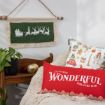 A cozy holiday scene with a red pillow reading “It’s the most wonderful time of the year,” a festive pillow, a green Santa wall hanging, a bench, a plant, and a gold ornament.