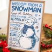 A framed sign titled “Wisdom from a Snowman” lists playful winter advice alongside a drawing of a snowman wearing a hat and scarf, surrounded by holiday decorations and greenery.