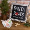A rustic sign reads “Santa Paws Is Coming to Town” with a paw print and Santa hat, next to a small pine tree, dog treats, a festive cloth, and a jar of peanuts on a wooden table.