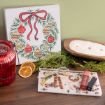 A festive scene with a painted Christmas wreath, red candle holder, dried orange slice, greenery, eyeglasses, a tray with makeup items, and a wooden bowl candle on a wooden surface.