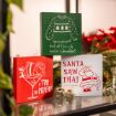 Three festive holiday signs: one with an ugly sweater, one with Santa in a martini glass, and one with Santa holding his belly. Christmas decorations and bells are in the background.