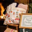 A cozy holiday scene with a blanket and pillow on a chair, a framed Merry Christmas sign, wrapped gifts, and a fireplace in the background.