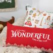 A festive scene with a red pillow that reads “It’s the most wonderful time of the year,” resting on a cream knit blanket, with a decorative holiday pillow featuring cars, gifts, and festive items in the background.