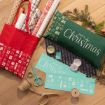 Christmas-themed gift wrapping supplies, a green “Merry Christmas” pillow, red tote bag, rolls of wrapping paper, scissors, twine, tape, and pine branches arranged on a wooden floor.