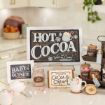A cozy kitchen hot cocoa station with a “Hot Cocoa” sign, jars of cocoa mix, peppermint sticks, mugs, and festive decorations, all arranged on a counter.