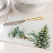 A gold-handled knife rests on a glass cutting board featuring a painted snowy log cabin scene surrounded by pine trees, placed on a marble countertop.
