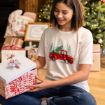A smiling person sits by a Christmas tree, holding a decorated gift box. They wear a white shirt with a red truck and trees on it. Wrapped presents are stacked in the background.