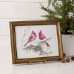 Framed artwork of two red cardinals perched on a snowy branch, displayed on a stand near a small potted evergreen with pinecones on a white surface.