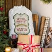 A large gift bag labeled “Special Delivery from Santa” sits on a chair next to wrapped presents, a spool of twine, rolls of wrapping paper, and a decorated Christmas tree with ornaments.