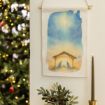 A watercolor nativity scene wall hanging shows Mary, Joseph, and baby Jesus under a stable with a bright star above. A candle and greenery are nearby, with a decorated Christmas tree blurred in the background.