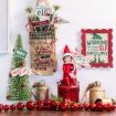 Festive holiday scene with an elf doll, mini Christmas tree, red ornaments, jingle bells, and colorful signs with cheerful messages like “I’m Back” and “Y’all Be Good Now.”.