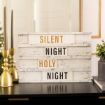 A decorative sign reads “SILENT NIGHT HOLY NIGHT” in gold and black letters, placed on a black table with gold candle holders, greenery, and a mirror in the background.