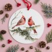 A round ornament with a painting of two red cardinals on snowy branches, surrounded by pinecones, fir sprigs, red berries, and small gold bells, all arranged on a pink background.