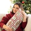 A woman in a red sweater smiles while hugging a festive holiday pillow on a couch, with a decorated Christmas tree in the background.