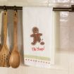 A white kitchen towel with a smiling gingerbread person missing part of one leg and the words “Oh Snap!” hangs on a rod next to wooden cooking utensils.