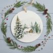 A round ornament with a snowy cabin and evergreen trees painting, surrounded by pine branches, pinecones, red berries, and wooden beads on a light blue wooden surface.