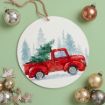 A round ornament featuring a red vintage truck carrying a Christmas tree, with snowy pine trees in the background. Festive ornaments and wooden decorations surround the ornament on a green surface.