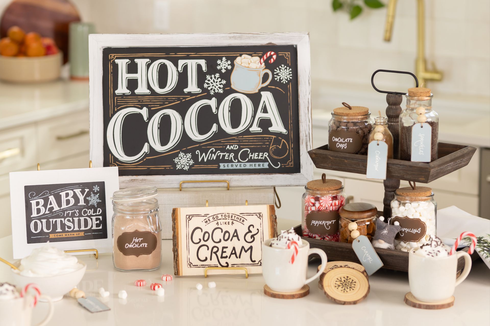 A kitchen counter displays a hot cocoa bar with mugs, labeled jars of toppings, a “Hot Cocoa” sign, and festive decorations. Marshmallows, chocolate chips, and peppermint sticks are visible among the treats.
