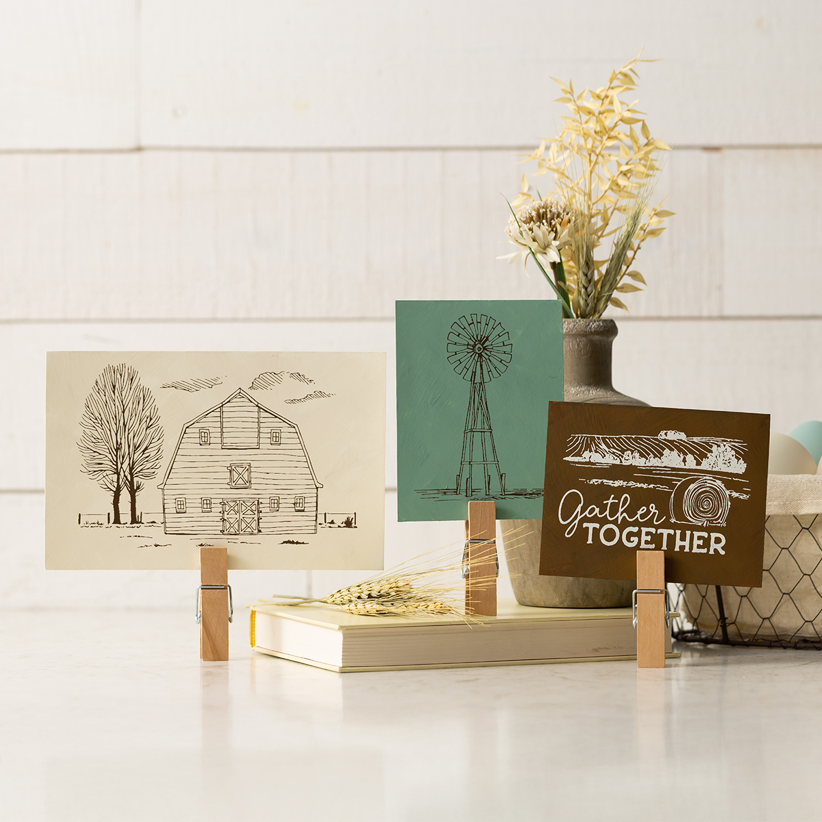 Three cards on wooden clips: one with a barn, one with a windmill, and one with the words “Gather Together.” A vase with dried flowers and a closed book are in the background, all set on a bright, neutral surface.
