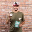A smiling man in a green shirt with a Mickey Mouse silhouette on the pocket holds a jar of black ink and a blue sheet with white Mickey Mouse shapes. He wears a red and white cap with a black Mickey Mouse silhouette.
