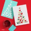 Two cards show Christmas trees made from Mickey Mouse head silhouettes; one card is turquoise, the other is white with red, green, and gold heads. A red paint jar and blue paint swatch sit on a red surface.