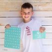Smiling child in a white shirt holds an alphabet stencil and a drawing stencil, standing against a wooden wall. The shirt features a cartoon character and the name “Oliver.”.