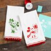 Two white holiday towels with red trim, featuring green Minnie Mouse and red Mickey Mouse designs holding gifts. Craft supplies and a teal stencil are nearby on a wooden surface.