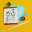 A holiday card featuring Mickey and Minnie Mouse, paint containers, a stencil, a scalloped border piece, and a blue eraser are arranged on a bright yellow surface. The card reads “Oh Golly more Holly!”.