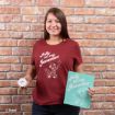 A smiling woman stands by a brick wall, holding a small jar and a mint-green sign that says “Jolly in my Jammies.” She wears a maroon T-shirt with the same phrase and a cartoon character.