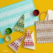 Holiday-themed Mickey Mouse print paper in blue, green, and red is shown alongside craft paint, a paper bag, and folded triangles for a DIY gift bag project on a yellow background.