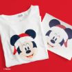 A white T-shirt with a print of Mickey Mouse wearing a Santa hat, placed next to a matching printed image, both on a red background.