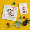 Holiday-themed flat lay with Mickey and Minnie Mouse sledding art, a card with colorful Mickey head silhouettes, a green tree-shaped item, small ornaments, fir branch, and a black container against a yellow background.