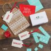 A brown gift bag with white Disney holiday patterns, red gift tags labeled “NOAH,” “EMILY,” and “GRACIE,” a red “OH JOY!” card, a “NOEL” card, stencils, and a red wax seal on a wooden table.