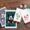 Three holiday-themed Disney hand towels with Mickey and Minnie Mouse designs are displayed on a wooden surface, next to red cord, wax seal, and crafting supplies. Disney copyright is visible in the corner.