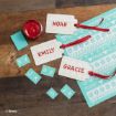 Three gift tags with “NOAH,” “EMILY,” and “GRACIE” are labeled in red. They are surrounded by blue letter stickers, a red ribbon, and a small red jar on a wood and tile surface. A blue Disney-themed sheet is nearby.