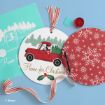 A round Christmas craft features Snoopy driving a red truck with a tree, “Home for Christmas” text, a red snowflake stencil, red paint, a ribbon, stencil sheet, and a paintbrush, all on a blue background.