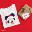 A white T-shirt with Mickey Mouse in a Santa hat next to a brown gift box tied with string, a pine sprig, and a name tag reading “Mickey! NOAH” on a red background.