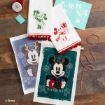 Disney-themed crafts featuring Mickey and Minnie Mouse: printed towels with festive designs, a green holiday banner reading “Yuletide Fun,” a red ink jar, stencil, string, and a wax seal on a wooden table.
