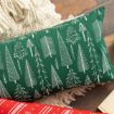 A green pillow with white illustrated Christmas trees sits on a wooden floor next to a cream-colored knit blanket. Part of a red fabric with white patterns is visible in the corner.