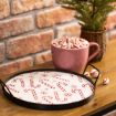 A pink mug filled with striped peppermint candies sits on a wooden table next to a tray decorated with candy cane and snowflake patterns. A small potted plant is in the background against a brick wall.