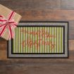 A wrapped gift with a red and white striped ribbon sits next to a doormat with green and white stripes and the words “merry christmas” written in red cursive. The floor is brown wood.