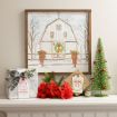 A holiday mantel with red flowers, wrapped gifts, a small green Christmas tree, and a framed barn illustration decorated with wreaths and red bows.