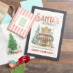 Flat lay of holiday-themed items on wood: framed “Santa’s Workshop” sign, tote with Christmas tree, striped card with “Ho Ho Ho,” tree-shaped cutting board, wrapped gifts, and a teal wax seal.