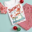 Flat lay of a festive pajama set with a white Snowman’s Creamery shirt, red checkered pants, Christmas ornaments, and a red holiday mug with “Let it Snow” text, all arranged on a light blue background.