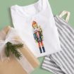 A folded white shirt with a nutcracker design, a brown gift box with a white ribbon and pine sprig, and a striped drawstring bag on a green background.